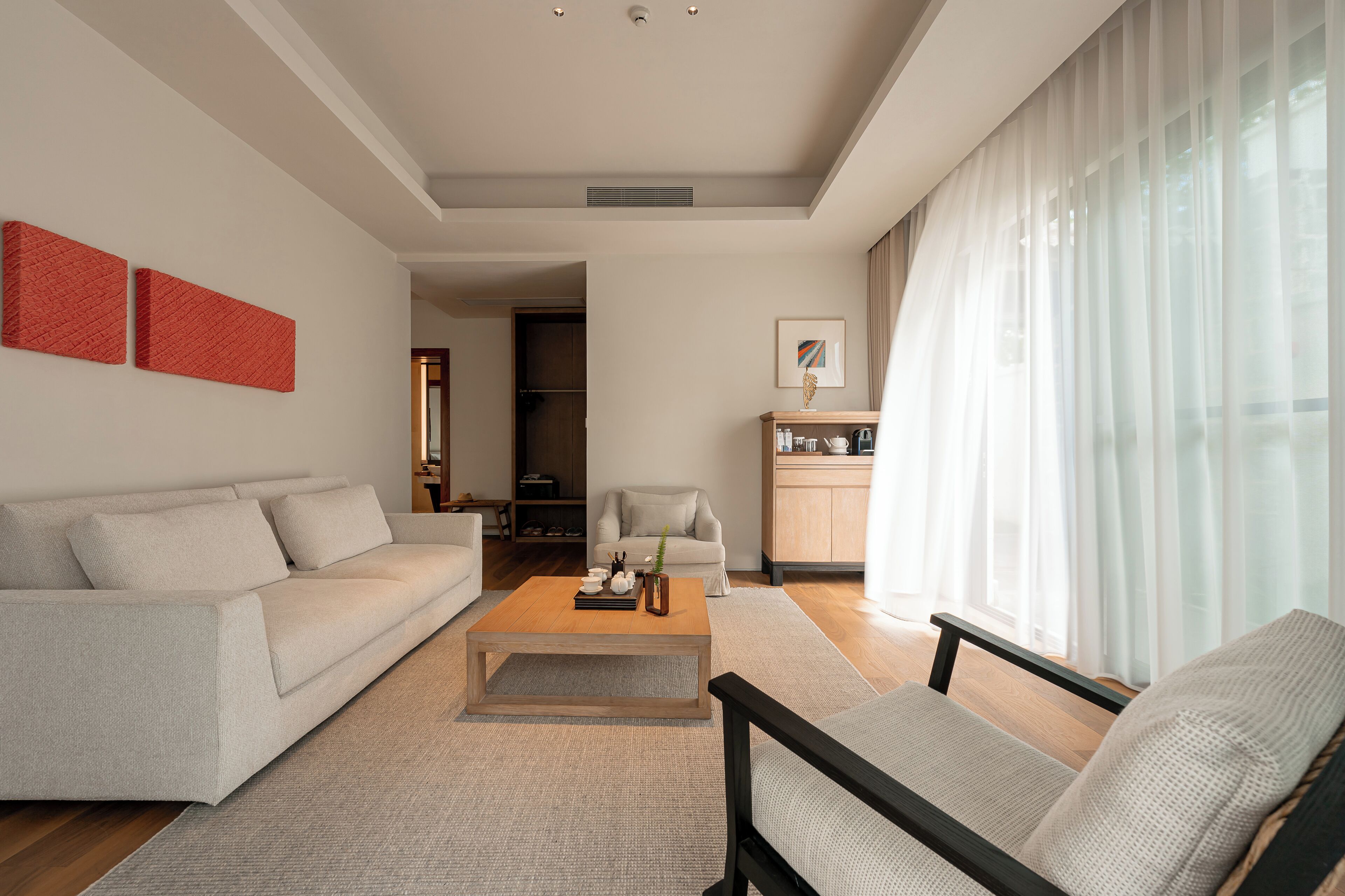 A modern living room with a sofa, armchair, coffee table, and a view of a kitchen and dining area through sheer curtains.