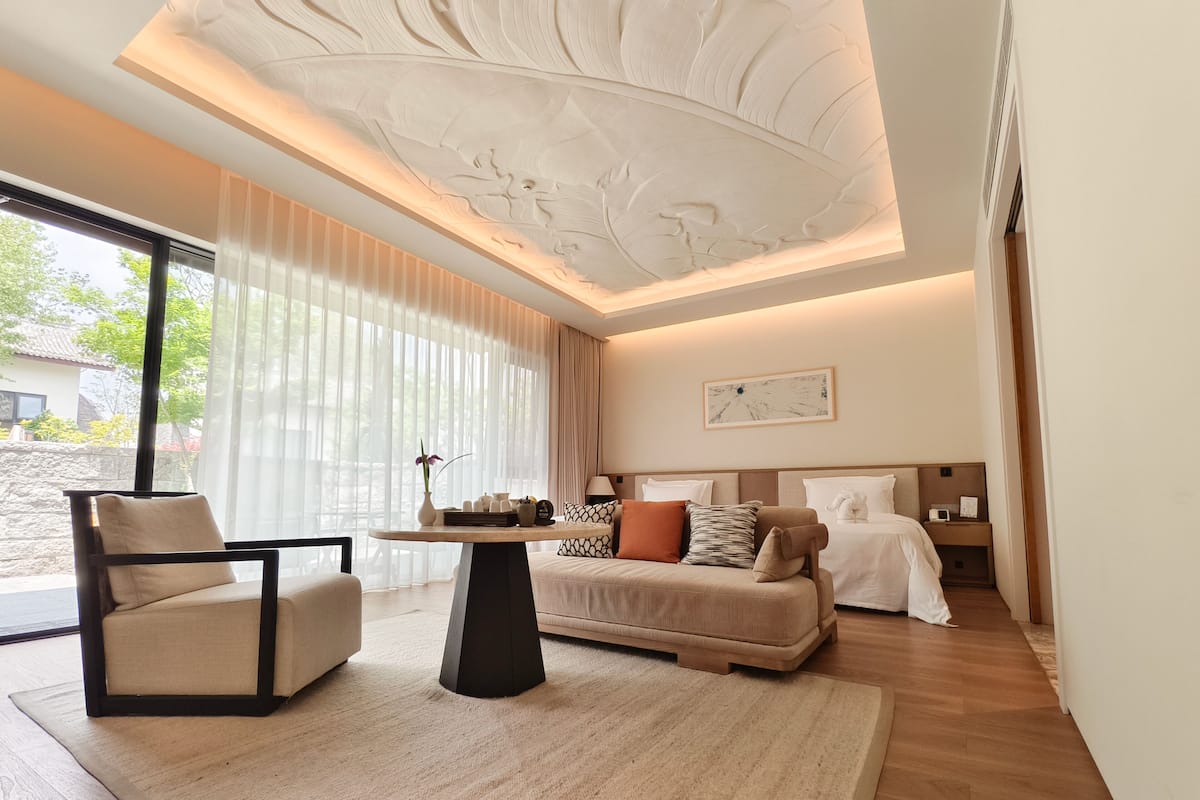 A modern hotel room with a sofa, a coffee table, a bed, and a large window with sheer curtains.