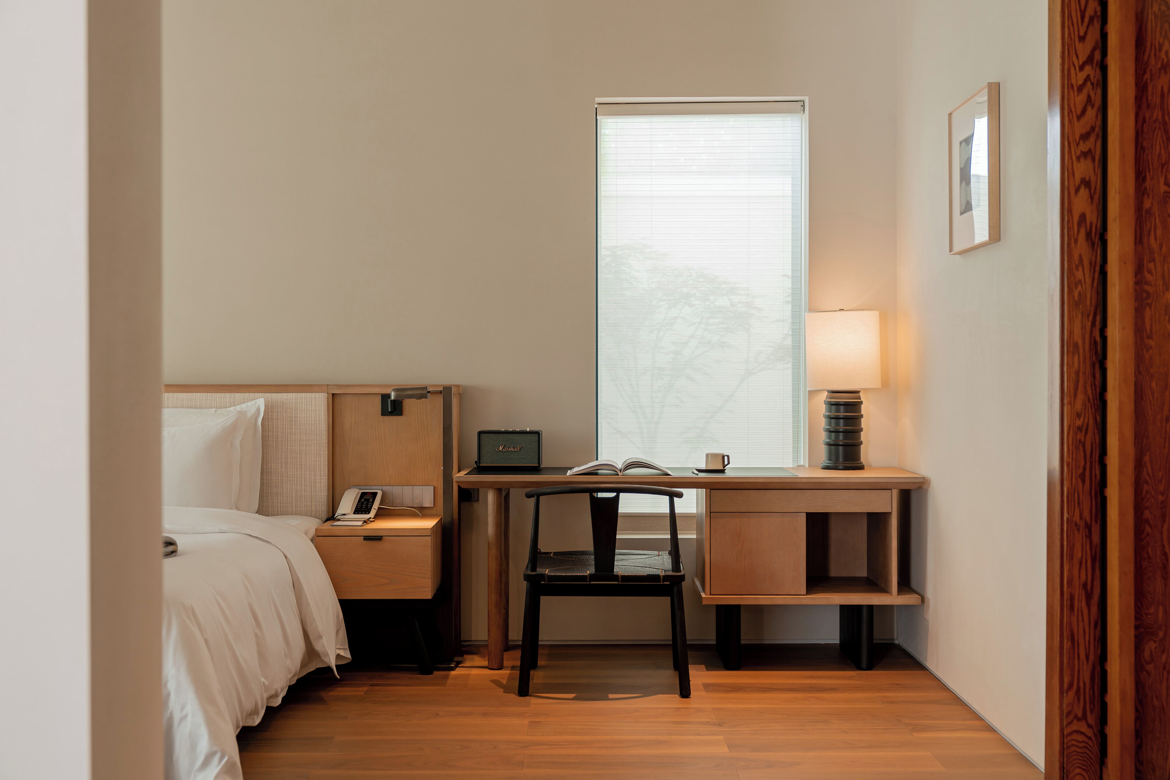 A hotel room with a bed, a desk with a lamp, a chair, and a window with blinds.