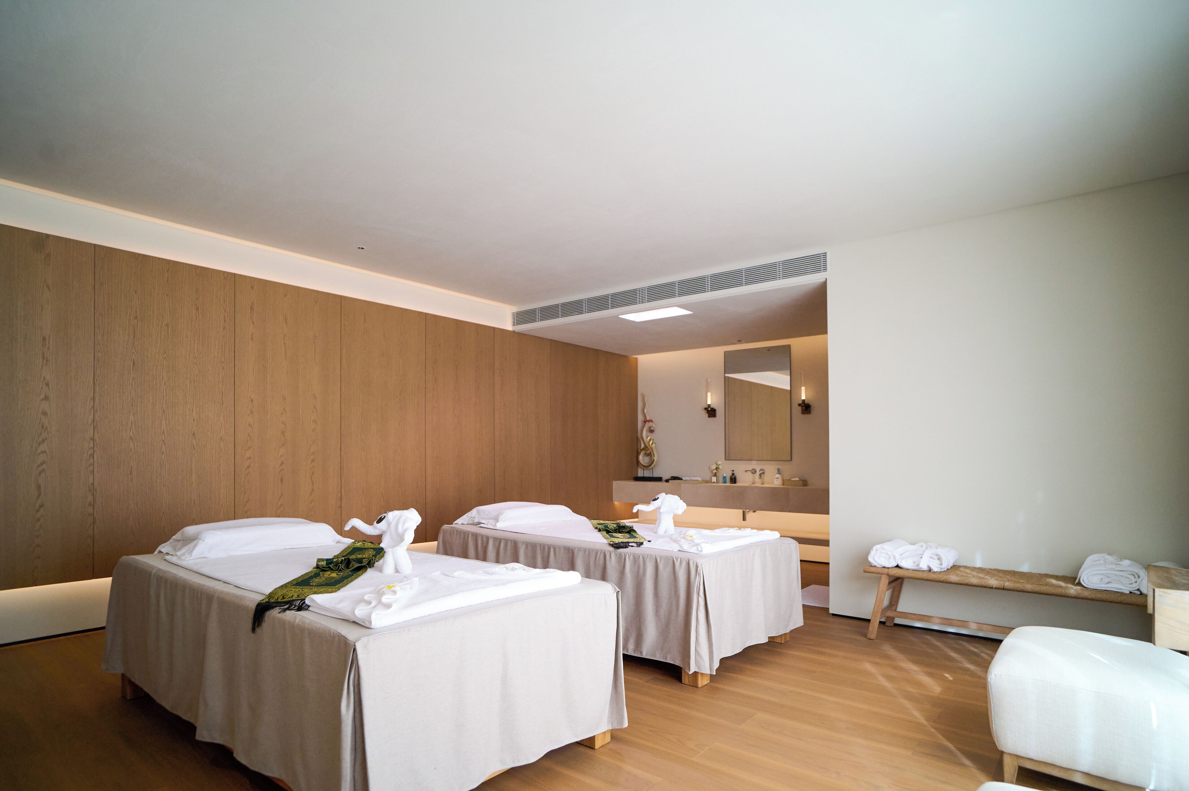A hotel room with two beds, a wooden wall, a mirror, and a bench with towels.