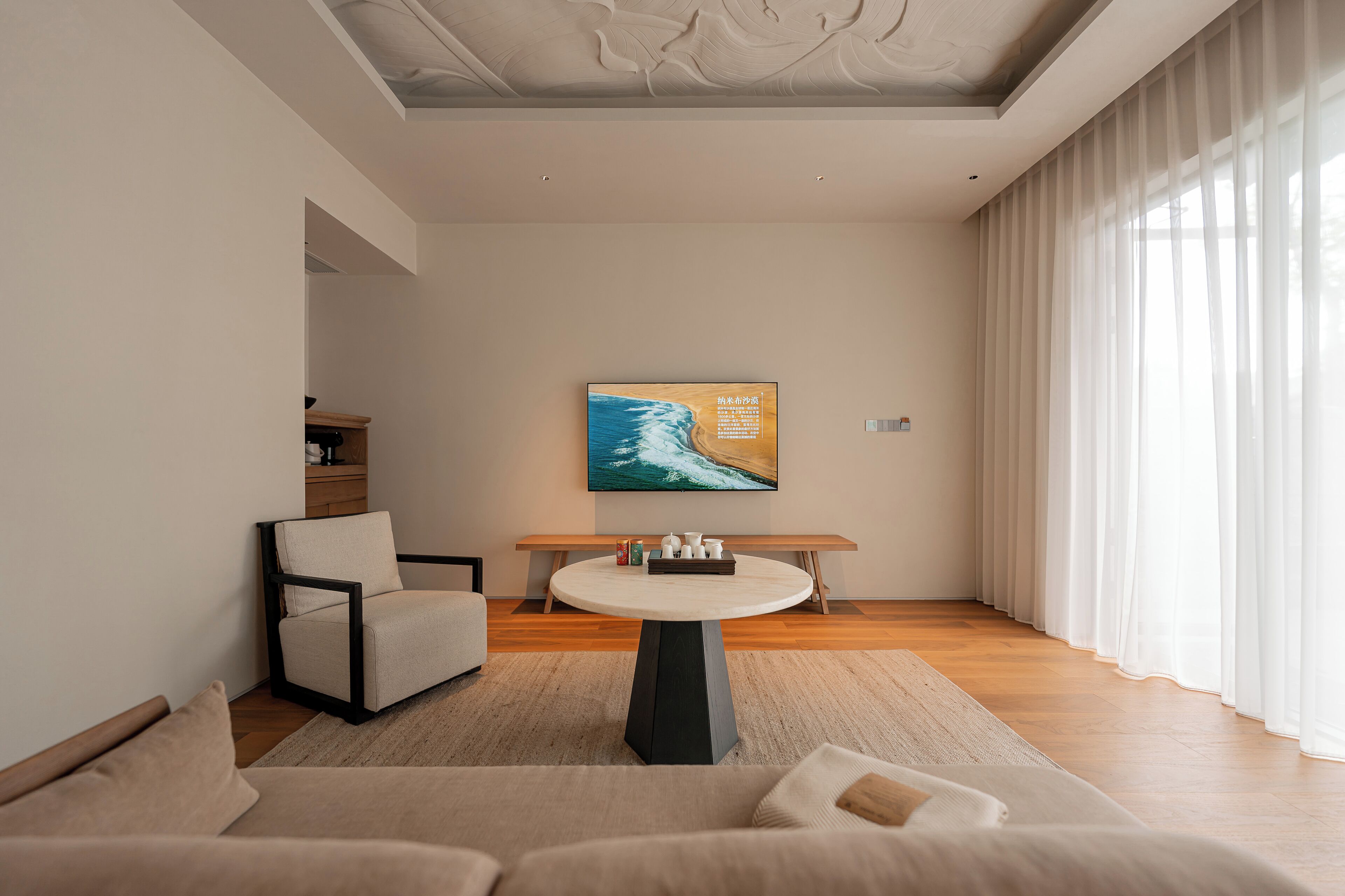 A modern living room with a round coffee table, a sofa, and a chair. There is a painting on the wall and a window with curtains.