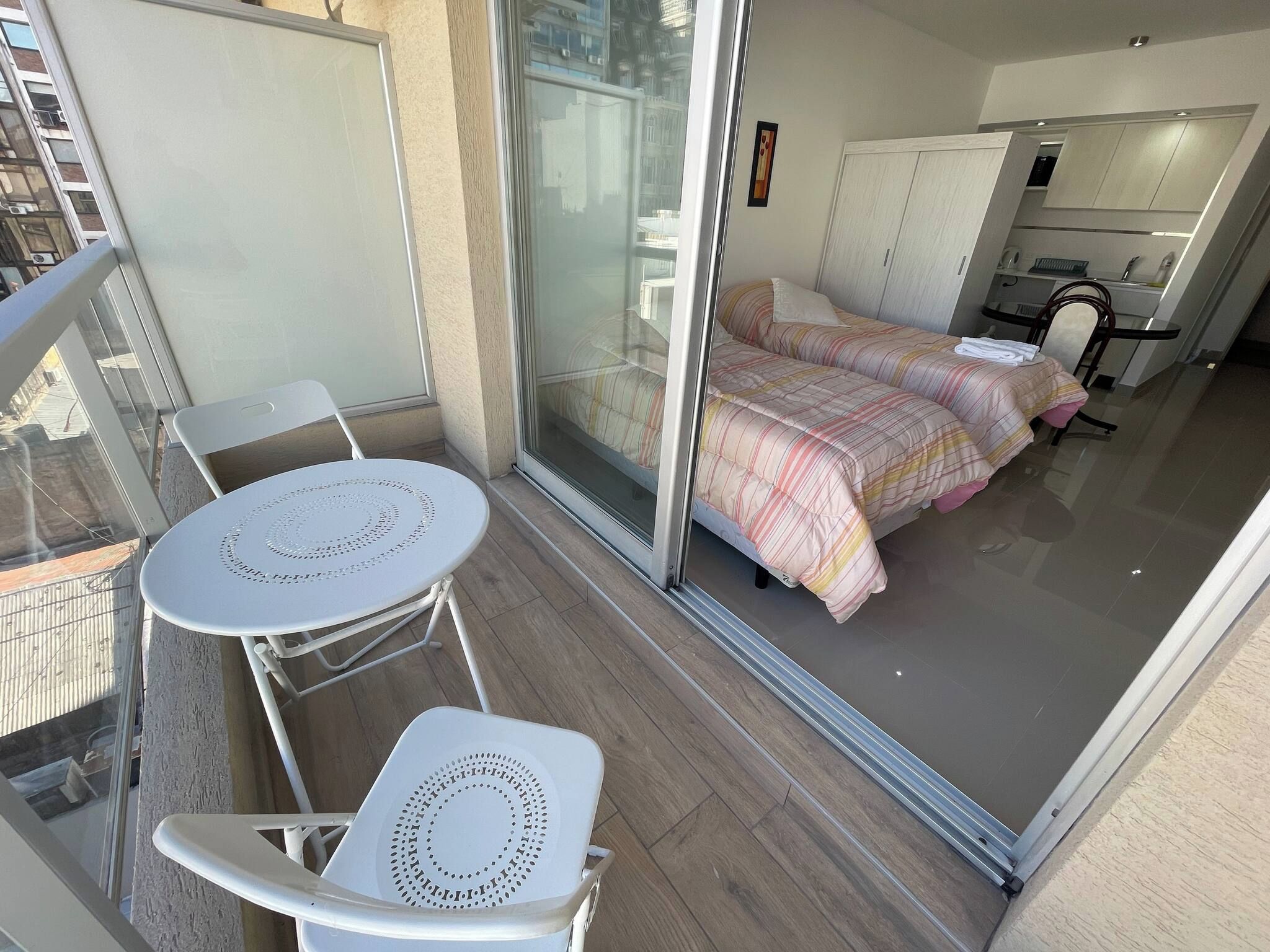 Un balcón con una mesa y sillas blancas, una cama con ropa de cama a rayas, y una zona de cocina con armarios y un lavabo.