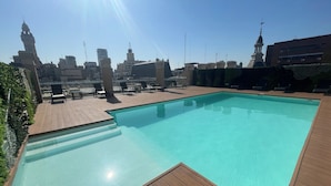 A rooftop swimming pool with wooden deck and lounge chairs, overlooking a cityscape with various buildings and a clear sky. - Studio In San Telmo With Balcony and Pool (Buenos Aires)