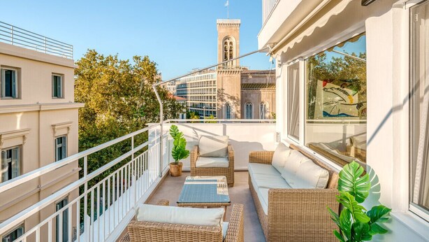 A balcony with wicker furniture, a potted plant, and a view of a building with a tower. - Achilleas 2-bedroom apartment (Athina)