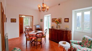 Un coin repas avec une table, des chaises et un lustre. On y trouve également un téléviseur posé sur un meuble, un vase avec des fleurs et une fenêtre donnant sur un beau paysage.