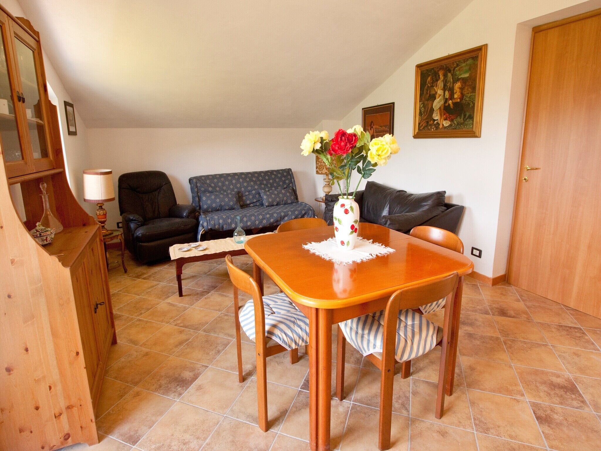 A dining area with a wooden table, chairs, a vase with flowers, a sofa, and a cabinet.