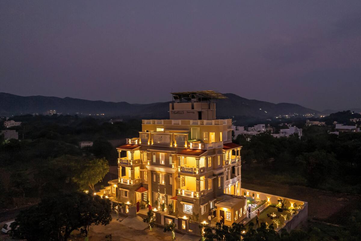 A multi-story hotel with a rooftop terrace and multiple balconies, surrounded by hills and trees.