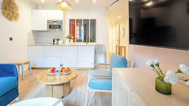Una sala de estar moderna con una mesa redonda, sofás azules, un televisor y una cocina con armarios blancos y un microondas.