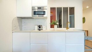 Una cocina moderna con armarios blancos, horno empotrado, lavamanos con grifo dorado y una ventana con cortinas.