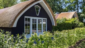 Ein kleines, dunkel gefärbtes Haus mit einem Strohdach und weißem Zierfach, umgeben von viel Grün.