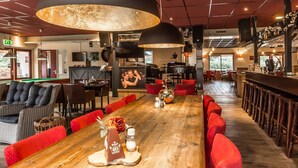 Ein großzügiger Barbereich mit hohen Decken, großen Pendelleuchten und einem langen Holztisch mit rot gepolsterten Stühlen.