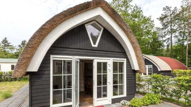 A small, dark-colored building with a thatched roof and a triangular gable. It has a large, open front door with glass panels and a small porch area. - Beautiful Cottage Near Giethoorn (De Bult)