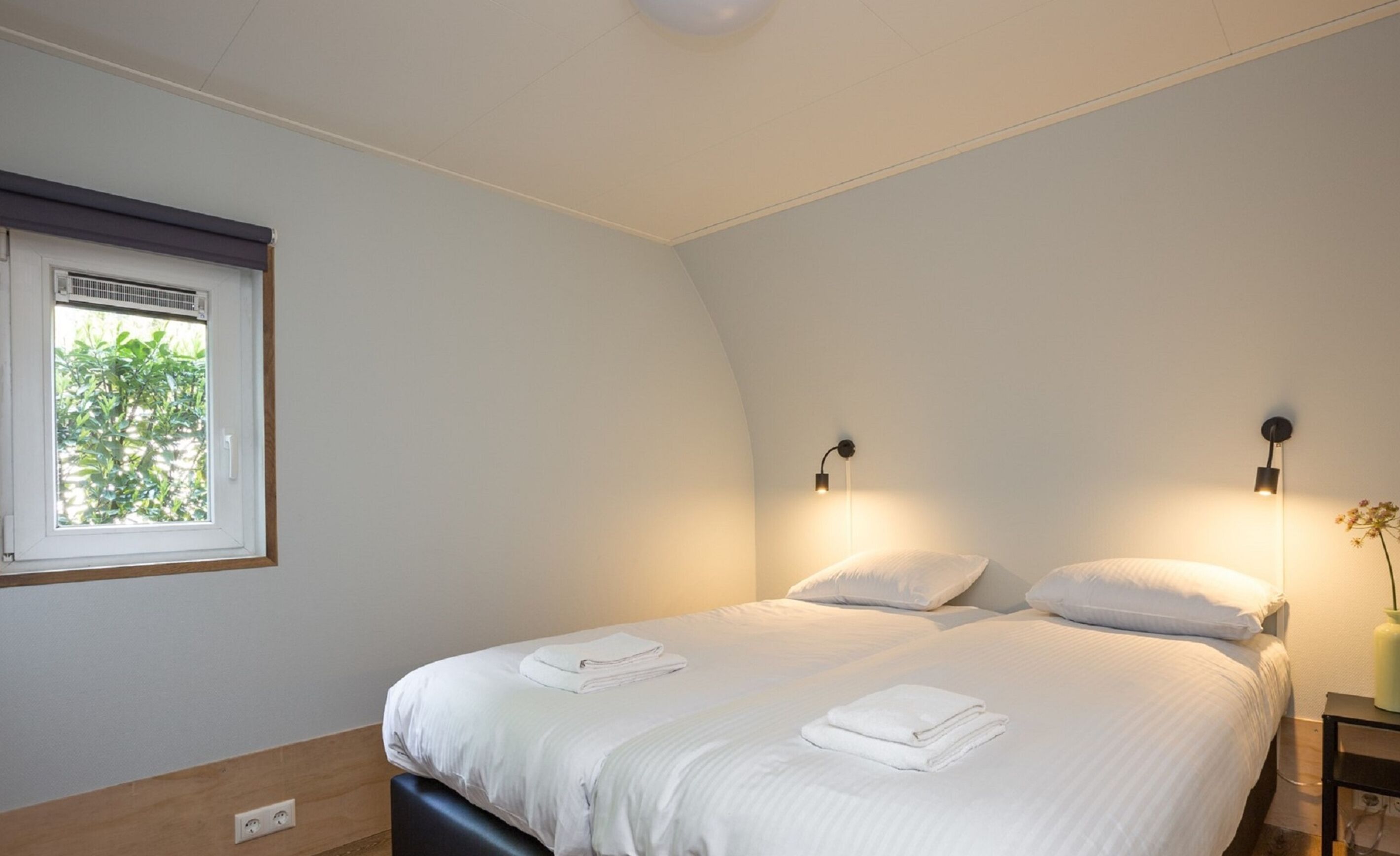 A bedroom with a bed, two bedside lamps, a window with blinds, and a nightstand with a vase.