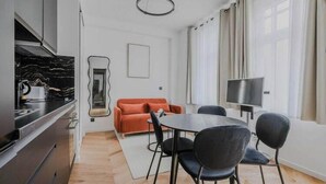 Une cuisine moderne avec un plan de travail noir et une cuisinière, un coin repas avec une table ronde et quatre chaises, un canapé et un téléviseur posé sur un meuble.