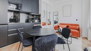 Une cuisine moderne avec des meubles de couleur sombre, une table de salle à manger ronde entourée de chaises noires et un canapé orange dans un salon lumineux.