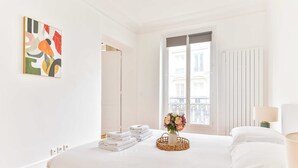 A hotel room with a bed, a window with a view of buildings, a wall-mounted artwork, and a bedside table with a lamp.