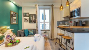 A modern hotel room with a bed, a sofa, a small dining area, a kitchen with a microwave, and a view of the city through a large window. - Lovely studio for 2 people - Sentier (Paris)