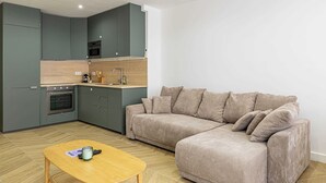 A modern kitchen with grey cabinets, a wooden countertop, and a built-in oven. A beige sofa with cushions is positioned against the wall, and a wooden coffee table is in the foreground.