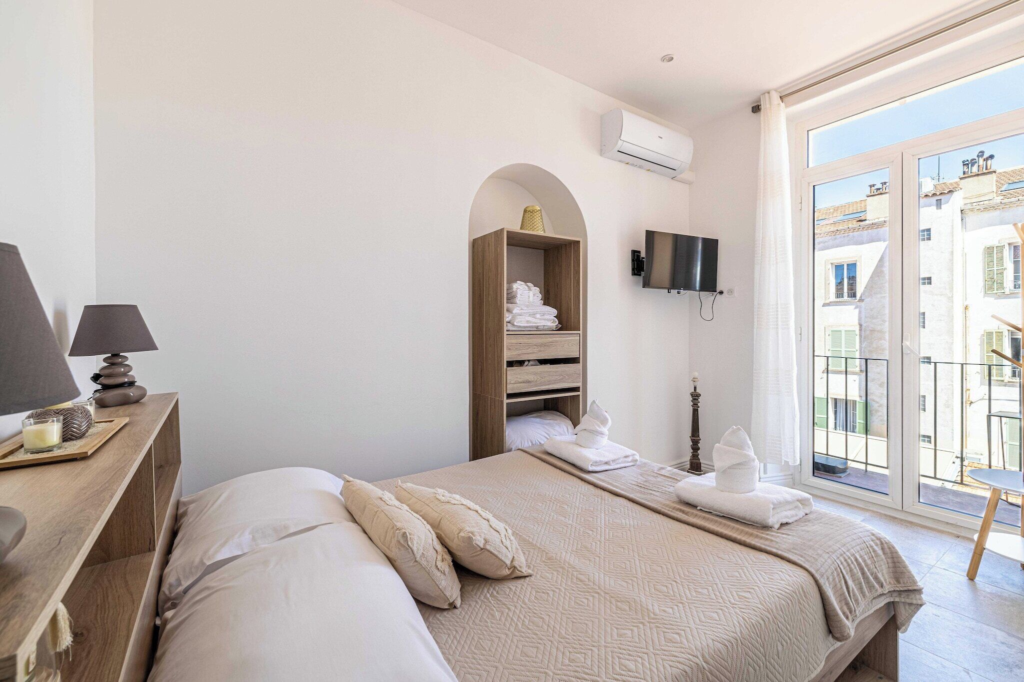 A hotel room with a bed, a wooden nightstand, a TV, and a balcony with a view of buildings.