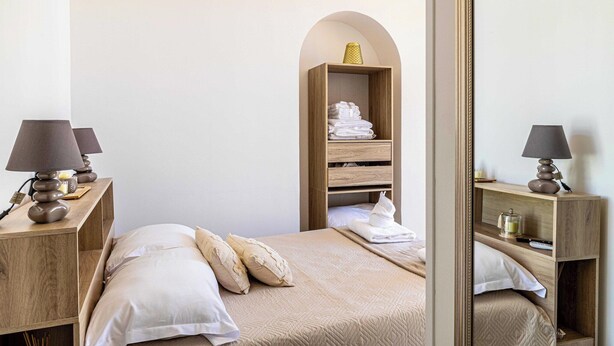 Une chambre à coucher bien rangée, avec un lit, deux tables de chevet dotées de lampes, une étagère en bois en forme d’arche où sont pliées des serviettes, et un miroir qui reflète la pièce.