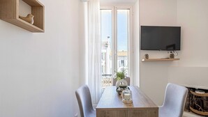 Un salon moderne avec une table à manger en bois, deux chaises grises, un téléviseur à écran plat fixé au mur et un balcon offrant une vue sur les immeubles.