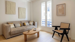 Una sala de estar moderna con un sofá gris, una mesa de centro de madera y una silla negra y marrón. Hay piezas de arte abstracto en las paredes y un cuadro enmarcado.
