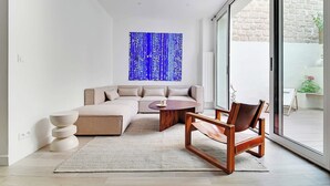A modern living room with a sectional sofa, a coffee table, and a chair. There is a large window with a view of a brick wall and greenery outside. - Charming aptt with patio - 2BD/4P - Monceau Park (Paris)