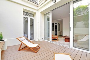 Une terrasse en bois avec un fauteuil de relaxation et une petite table, une porte-fenêtre coulissante donnant sur un espace intérieur comprenant un canapé et une table basse.