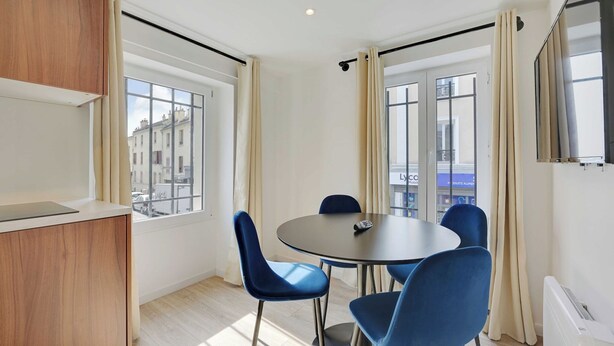 Ein modernes Zimmer mit einem runden Tisch, blauen Stühlen, einer Holzkabine und großen Fenstern mit Vorhängen.