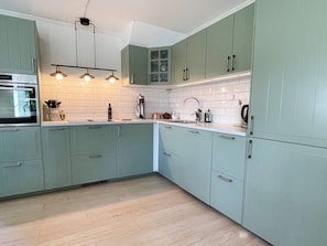 Een keuken met lichtgroene kastjes, een witte betegelde achterwand, een roestvrijstalen afzuigkap en een moderne hanglamp.
