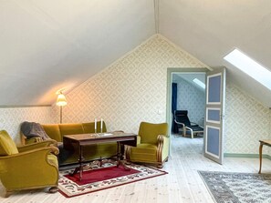 Een kamer met een schuine kap, een tafel met twee kaarsen, een bank, twee fauteuils en een kamerbreed tapijt met een patroon.
