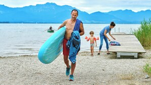 A person carrying a surfboard and a towel, walking on a pebbled beach with a wooden pier and mountains in the background. - Vacation apartment Kampenwand, 1-4pers., 2 sep. SZ, 1ST FLOOR + 2ND FLOOR (Chieming)