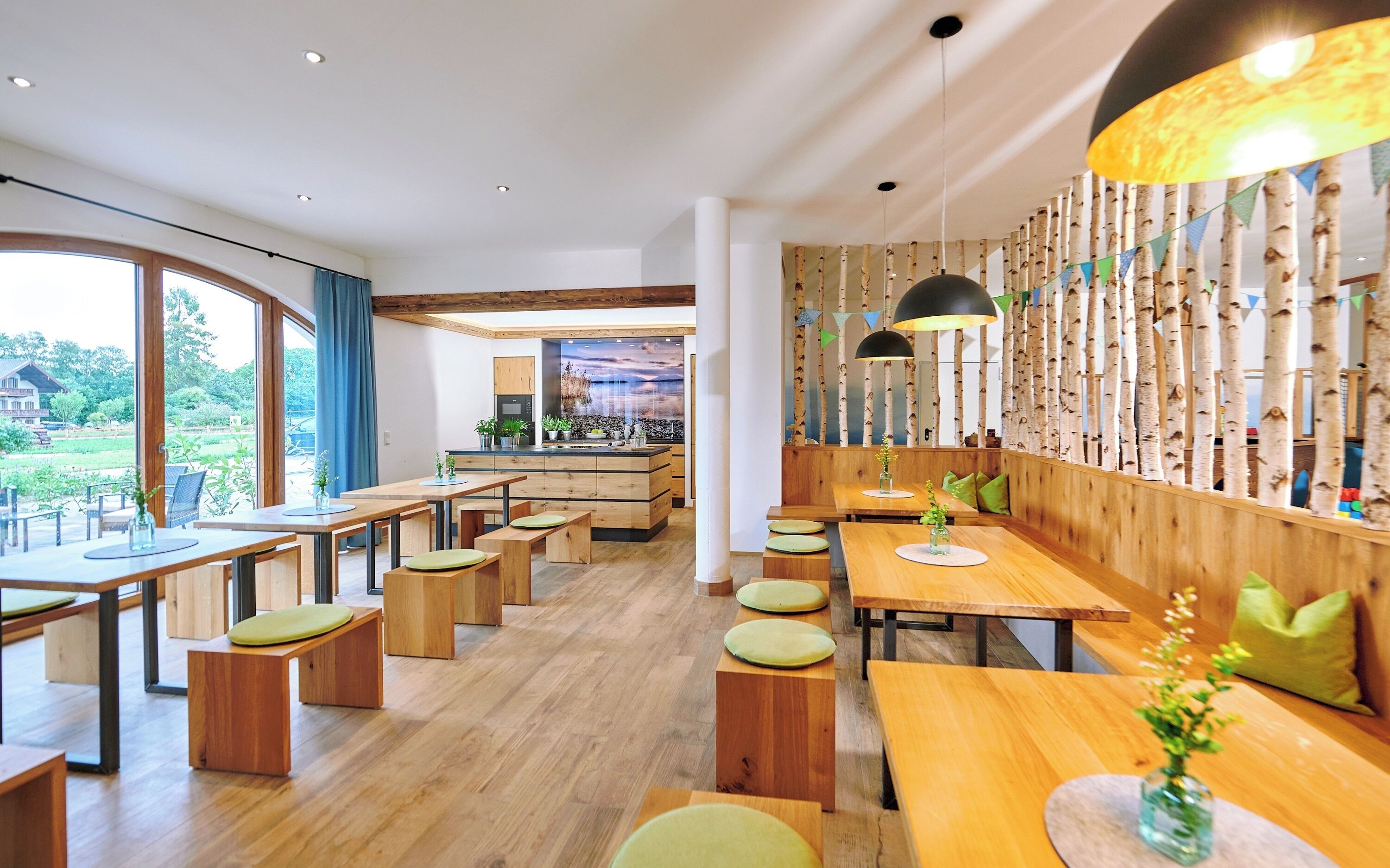 A modern restaurant with wooden tables and benches, a kitchen area, and large windows offering a view of the outdoors.