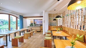 A modern restaurant with wooden tables and benches, a kitchen area, and large windows offering a view of the outdoors. - Vacation apartment Kampenwand, 1-4pers., 2 sep. SZ, 1ST FLOOR + 2ND FLOOR (Chieming)