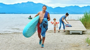 A person carrying a surfboard and a towel, walking on a pebbled beach with a wooden pier and mountains in the background. - Vacation apartment Hochfelln, 1-6pers., 2 sep. 1st bedroom, 1st floor, east-facing balcony with lake & distant view (Chieming)