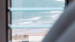 A view through a window with horizontal blinds, revealing a beach with waves and a sandy shore. - Retreat by the Sea – Coastal Escape with Cozy Spa & Panoramic Ocean Sunset Views (Gerroa)