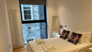 A modern hotel room with a large bed, two pillows, a nightstand, a lamp, and a view of a stone building through a window. - Stunning Park View Quartermile Apartment Close Proximity to Castle & University (Edinburgh)