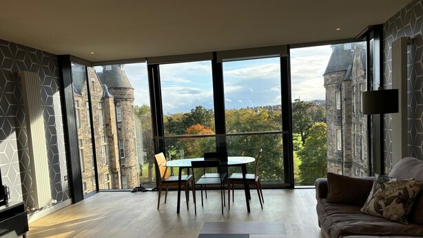 A spacious room with a large window offering a view of a historic building and trees. - Stunning Park View Quartermile Apartment Close Proximity to Castle & University (Edinburgh)