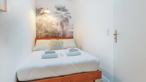 Una cama bien hecha con sábanas blancas y dos toallas dobladas, cabecera de madera, lámpara fijada a la pared y un cuadro con palmeras.