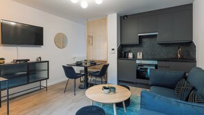Una sala de estar moderna con un televisor de pantalla plana, un comedor con mesa y sillas, y una kitchenette con armarios oscuros y una estufa.