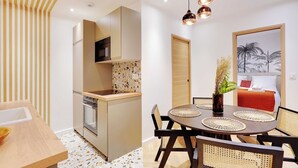 A modern kitchen with a microwave, oven, and a backsplash with a mosaic pattern. There is a dining area with a round table, chairs, and a vase with plants. A bed with a red blanket and a framed picture of palm trees is visible in the background. - Magnificent Apartment 1BR/4P - Arc de Triomphe (Neuilly-sur-Seine)