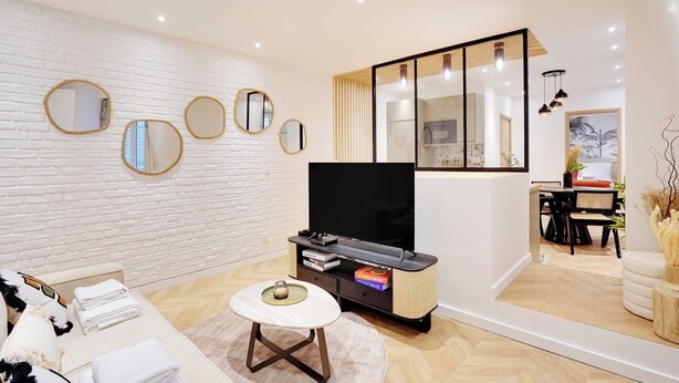 A modern living room with a white brick wall, a flat-screen TV, and a dining area with a black table and chairs. - Magnificent Apartment 1BR/4P - Arc de Triomphe (Neuilly-sur-Seine)