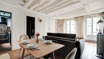 Un coin repas avec une table en bois dressée pour quatre personnes, un canapé noir et une cuisine visible au fond.