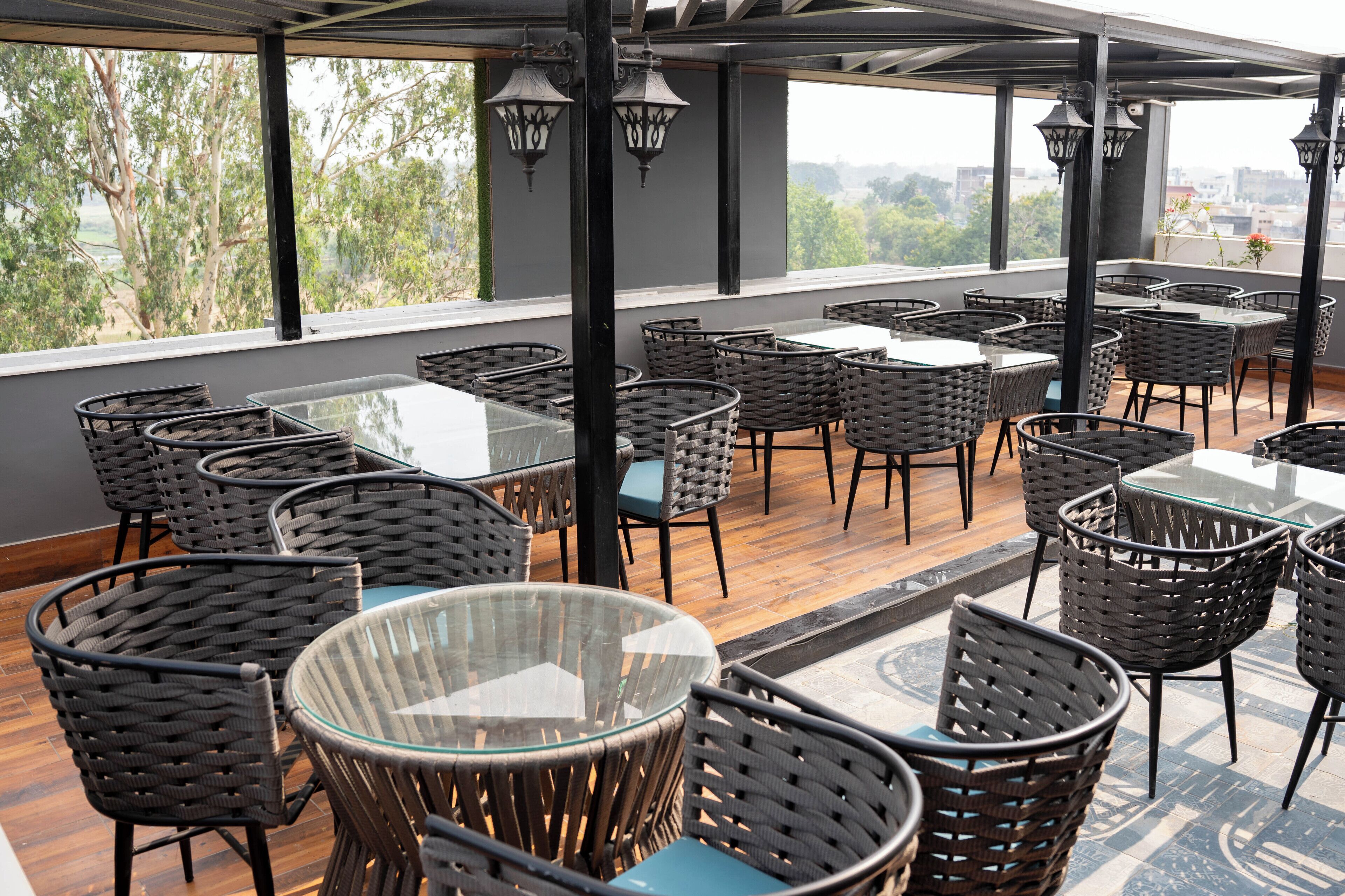 Un espace extérieur aménagé avec des chaises en osier et des tables en verre, de grandes fenêtres offrant une vue sur les arbres et les bâtiments.