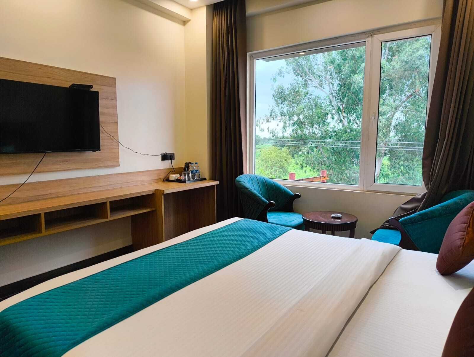 Habitación de hotel con cama, televisión montada en la pared, una mesita, una silla y ventana con vistas a árboles.
