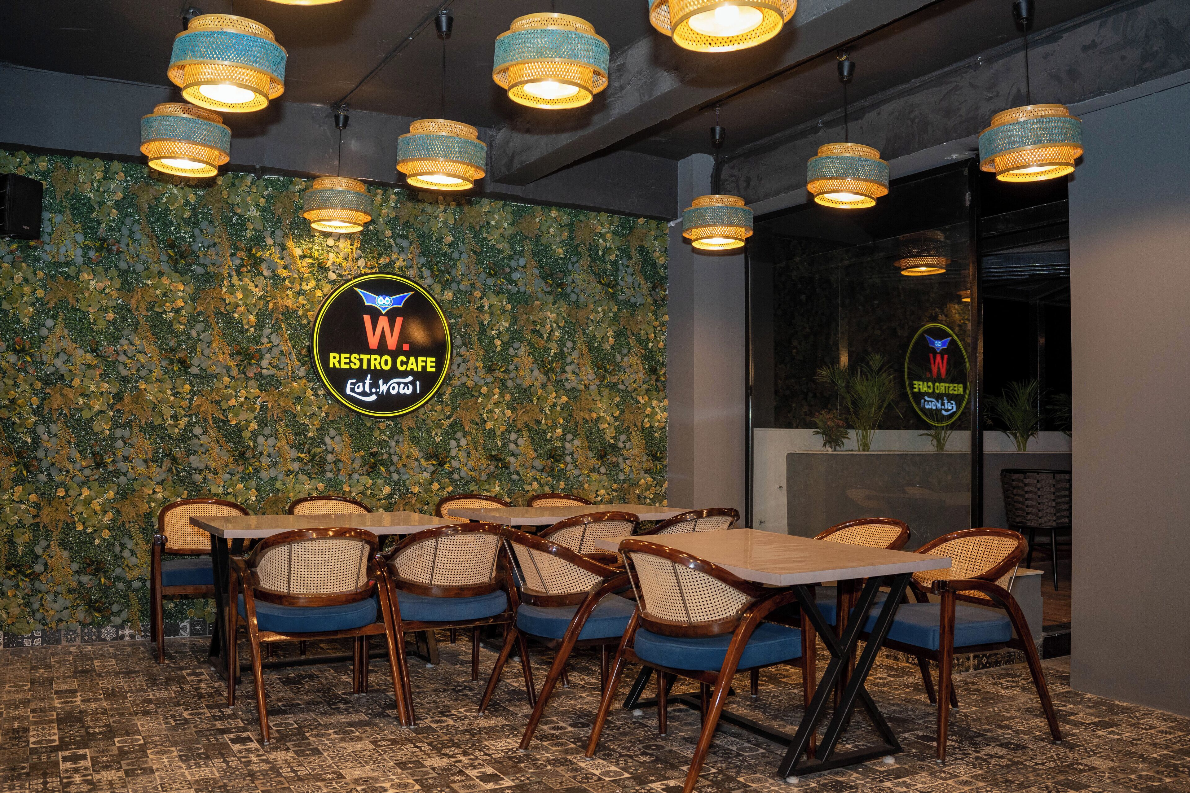 A restaurant with a green mosaic wall, wooden tables, and wicker chairs.