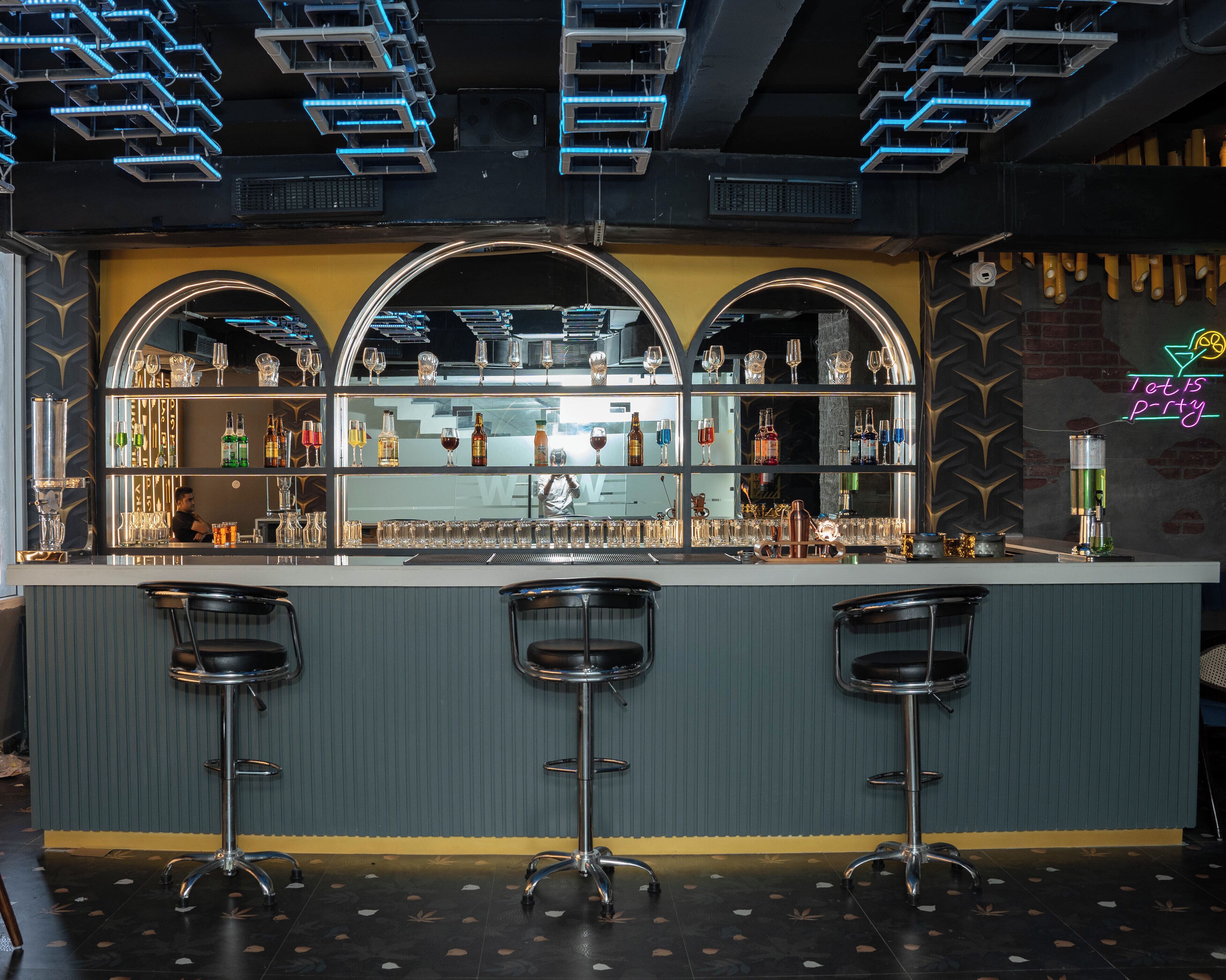 Un bar con tres taburetes, estantes llenos de botellas y vasos, y un letrero de neón.