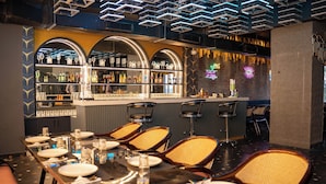 A restaurant with a long dining table set for a meal, bar area with shelves of bottles, and a neon sign on the wall. - Hotel W Grand - A Boutique Hotel (Haridwar)
