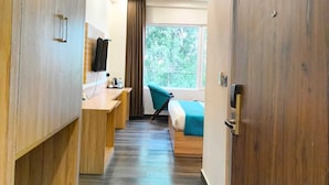 A hotel room with a wooden wardrobe, a desk with a chair, a bed with a blue blanket, and a window with a view of trees. - Hotel W Grand - A Boutique Hotel (Haridwar)