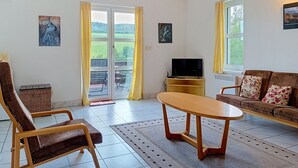 A living room with a sofa, a chair, a table, a TV, and a view of a green landscape through a glass door. - Green Valley Park Stárkov 13 by Interhome (Stárkov)